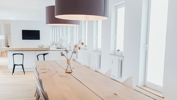 Et langt træbord med sorte metalben, en mørk hængende lampe og en vase med tørrede blomster.