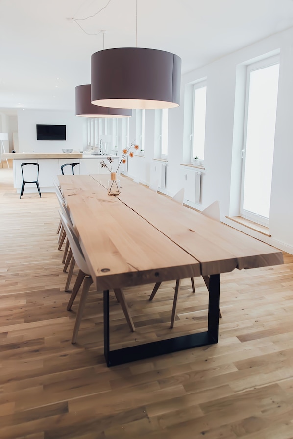 A long wooden table with black metal legs, a dark hanging light fixture, and a vase with dried flowers.