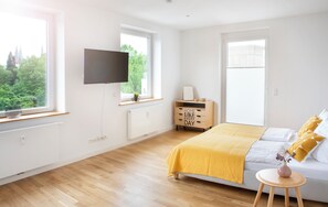 A bright bedroom with a bed, a TV, a dresser, and a window with a view of trees.