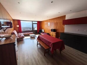 Ein modernes Hotelzimmer mit Holzboden, einer Couch, einem Esstisch mit roter Tischdecke, einem Fernseher und Blick auf die Berge.