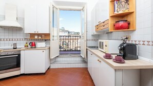 Μια μικρή κουζίνα με λευκά ντουλάπια, εντοιχισμένο φούρνο, φούρνο μικροκυμάτων και τοστιέρα. Υπάρχει ένα μικρό παράθυρο που οδηγεί σε ένα μπαλκόνι με θέα στα κτίρια.