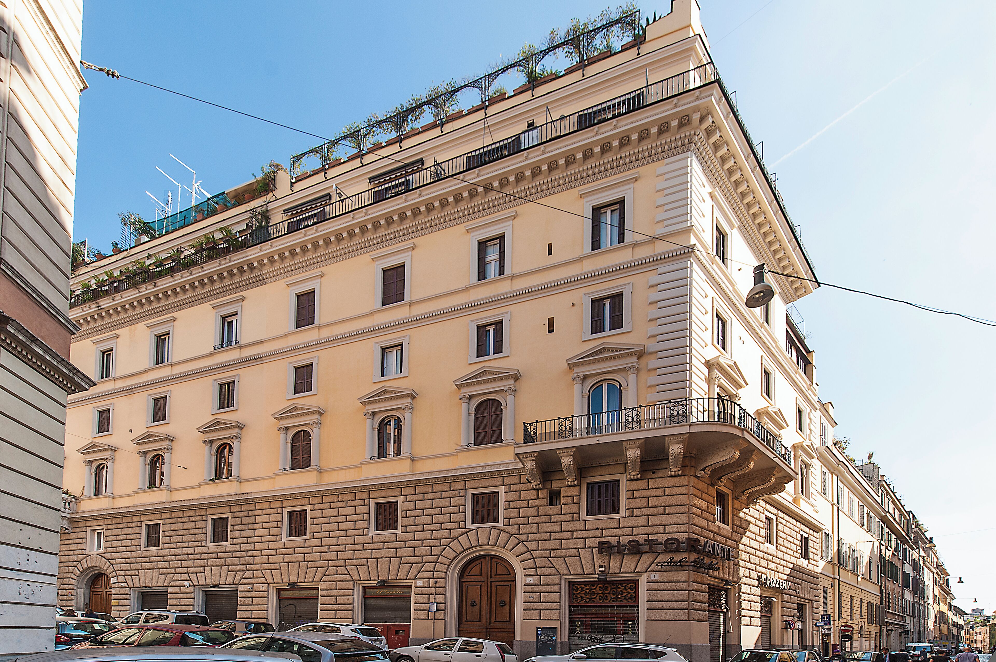 Ein mehrstöckiges Gebäude im klassischen Stil, mit einem Eingangsbereich mit großem Rundbogen und zahlreichen Fenstern auf jeder Etage, sowie einem Balkon mit Metallgeländer in den oberen Stockwerken.