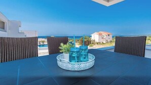 Ein Esstisch mit Getränken auf einem Tablett, ein Korbstuhl und Blick auf das Meer und Gebäude.
