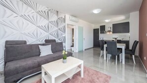 Ein modernes Wohnzimmer mit grauem Sofa, einem weißen Couchtisch und einer Wand mit Muster. Es gibt einen Essbereich mit Tisch und Stühlen sowie eine Küchenzeile mit Fernseher und Schränken.