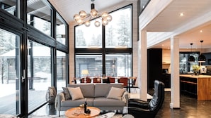 A spacious living area with a large gray sofa, a black leather chair, and a wooden coffee table. A dining area with a wooden table and chairs is visible in the background. - High-Quality 4BR Villa in Ruka - Private Hot Tub & Sauna (Kuusamo)