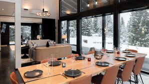 A modern dining area with a large table set for six, large windows offering a snowy landscape view, and a cozy seating area with a sofa and lamp. - High-Quality 4BR Villa in Ruka - Private Hot Tub & Sauna (Kuusamo)