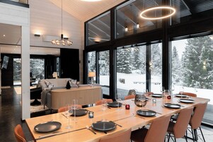 A modern dining area with a large table set for six, large windows offering a snowy landscape view, and a cozy seating area with a sofa and lamp.