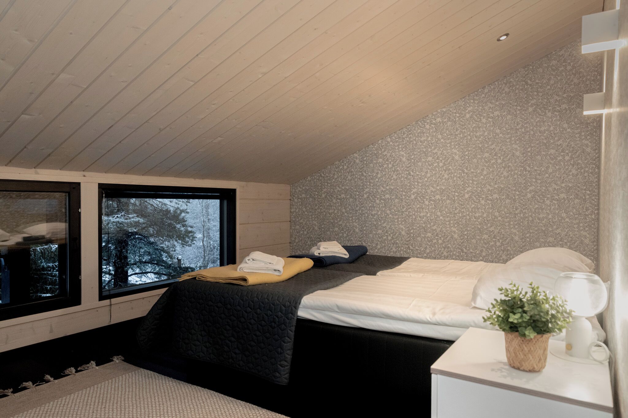 A modern bedroom with a large bed, a bedside table with a lamp, and a window with a view of trees.
