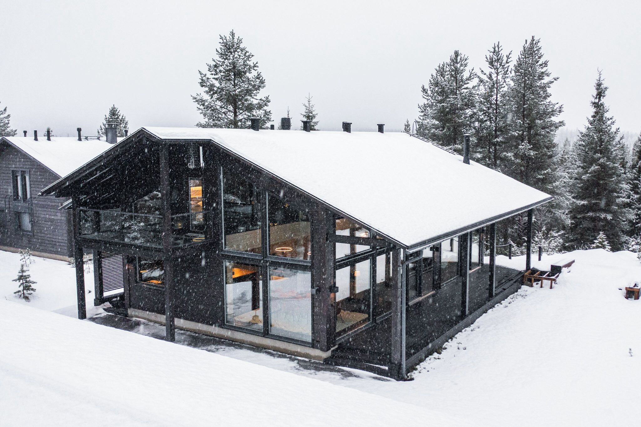 A modern wooden cabin with large glass windows, surrounded by snow-covered trees.
