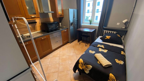 A compact hotel room with a kitchenette, a bed with a butterfly-themed bedspread, a small desk, and a window with a view of buildings. - Fantastic Inn Warsaw - CH16 (Warsaw)