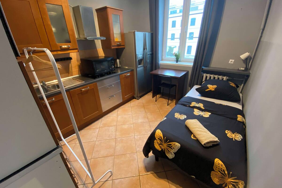 A compact hotel room with a kitchenette, a bed with a butterfly-themed bedspread, a small desk, and a window with a view of buildings.