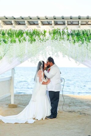 A couple kissing under a canopy with greenery overhead, a beach in the background, and a white structure with a roof. - Sunshine Cabanas At Ocean View (Dangriga)