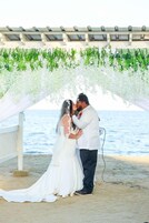 Una pareja besándose bajo una estructura cubierta de vegetación, con una playa al fondo y una construcción blanca con techo.