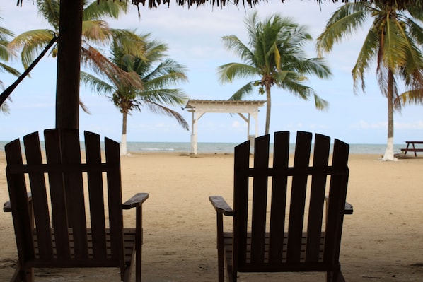 Two wooden chairs facing a beach with palm trees and a gazebo.