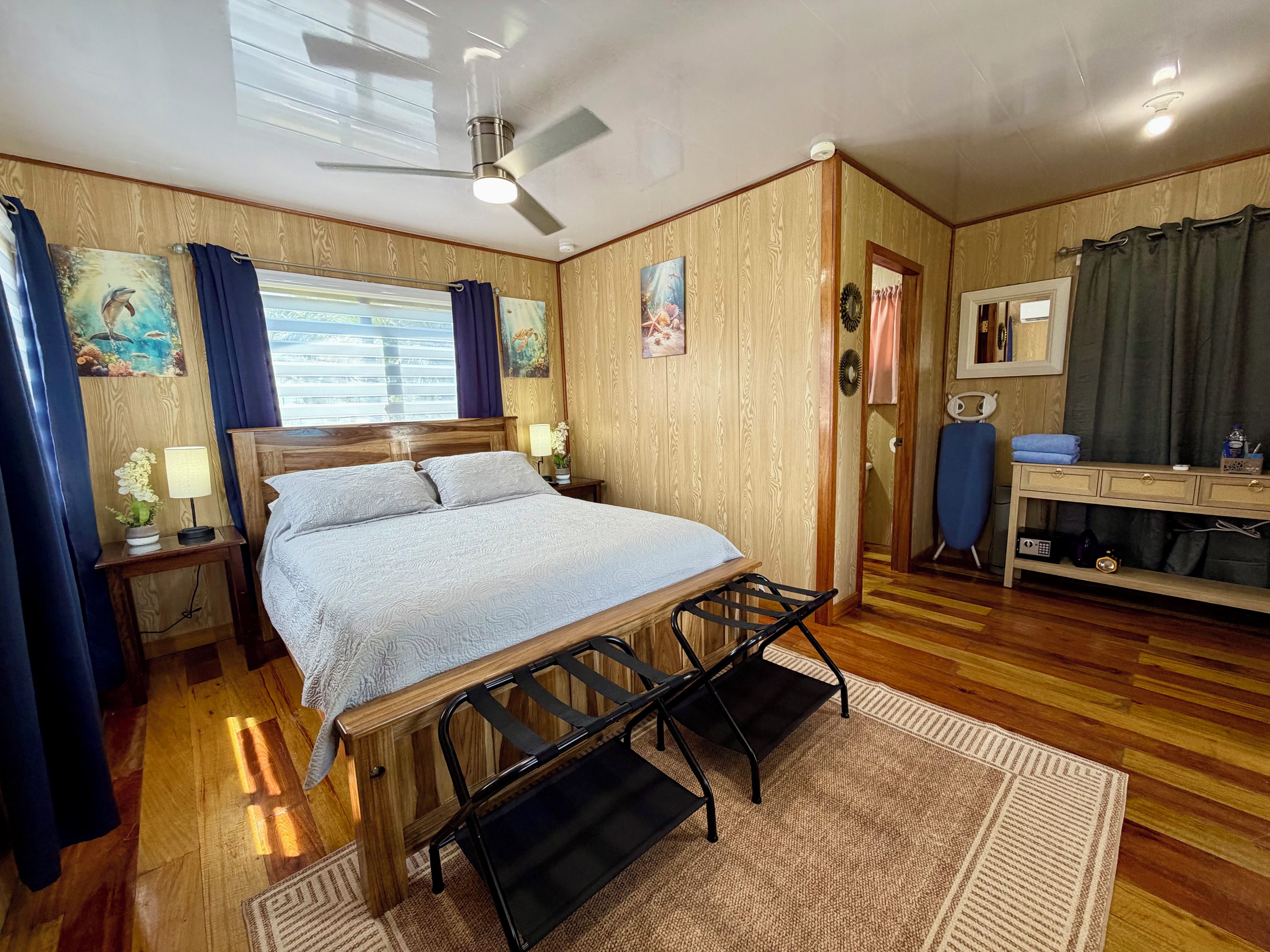 A bedroom with a bed, bedside tables, a desk, a chair, a ceiling fan, and a window with curtains.