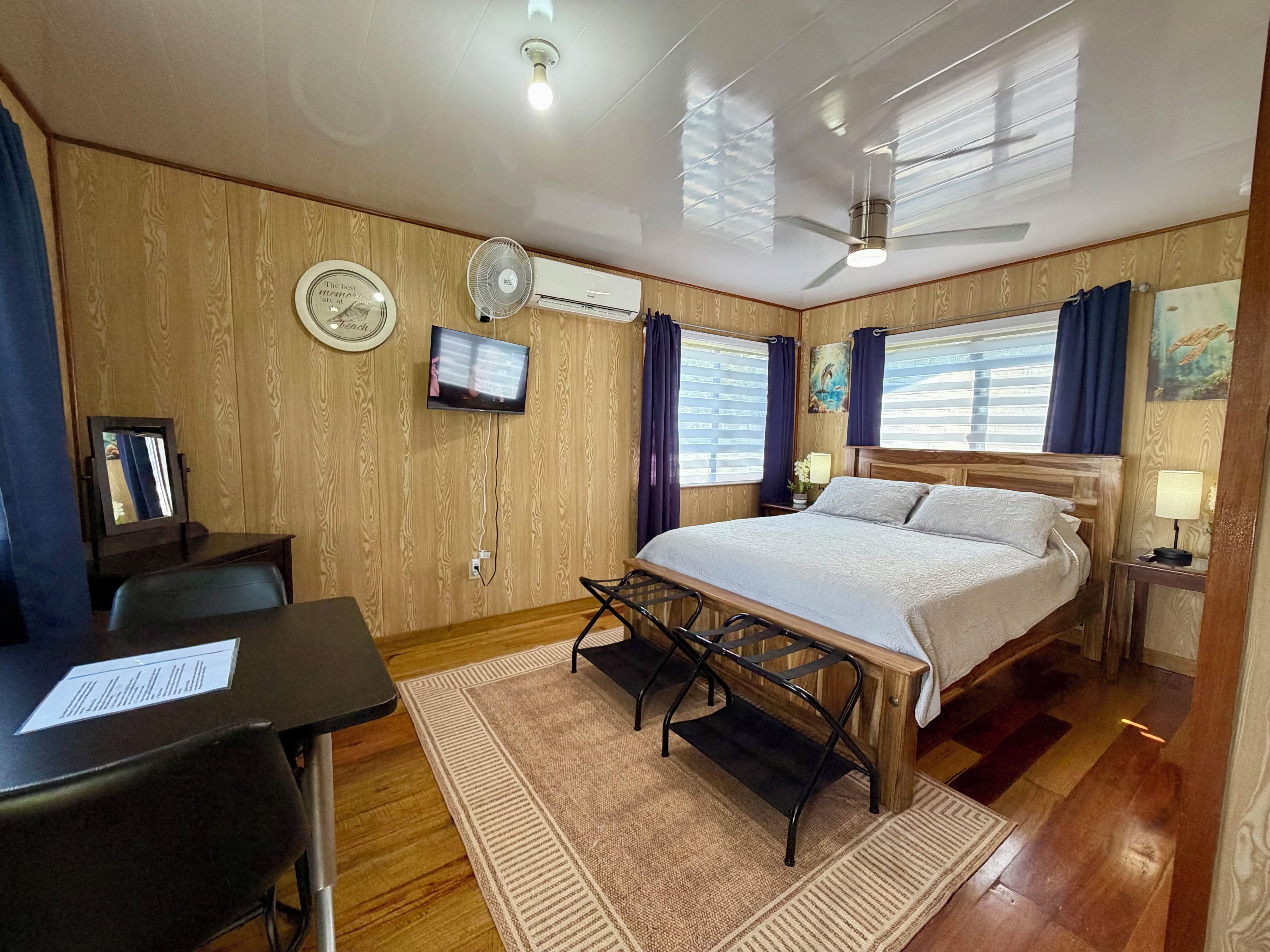 Une chambre d’hôtel équipée d’un lit, d’un bureau, d’une chaise, d’une télévision, d’un ventilateur de plafond et de murs en bois.