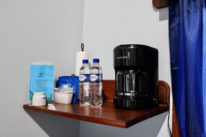 A small wooden table with a coffee maker, two water bottles, a cup, a tissue roll, and a blue bag.
