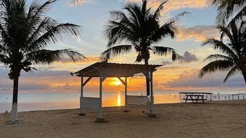 Une plage avec un kiosque, des palmiers et un coucher de soleil.