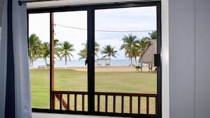A room with a large window overlooking a beach with palm trees and a thatched-roof structure. - Sunshine Cabanas At Ocean View (Dangriga)
