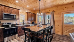 A wooden cabin kitchen with a central island, stainless steel appliances, and a large window with curtains. - Quiet Cabin Near Pkwy | Hot Tub | Game Room (Pigeon Forge)