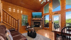 Interior de una cabaña de madera con chimenea de piedra, un sofá de cuero, una mesa de comedor y vistas a través de la puerta.