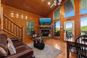 A wooden cabin interior with a stone fireplace, a leather sofa, a dining table, and a view through the door.