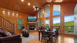 Interior de una cabaña de madera con chimenea, televisor, zona de comedor y una escalera.
