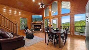 Interior de una cabaña de madera con chimenea, televisor, zona de comedor y una escalera.