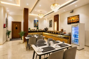 A hotel dining area with tables set for guests, a buffet counter, and a refrigerator stocked with beverages. - Hotel Grand Shalimar by JMD Group (New Delhi)