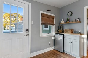 Une cuisine avec une porte blanche, une fenêtre avec un store marron, une horloge au mur et une étagère ornée d’objets décoratifs.