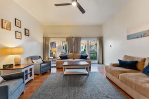 A living room with a sofa, a coffee table, and a lamp. There is a ceiling fan and a window with curtains.