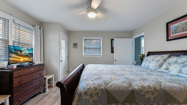 Un dormitorio con cama, cómoda, televisor, ventilador de techo y ventana con persianas.