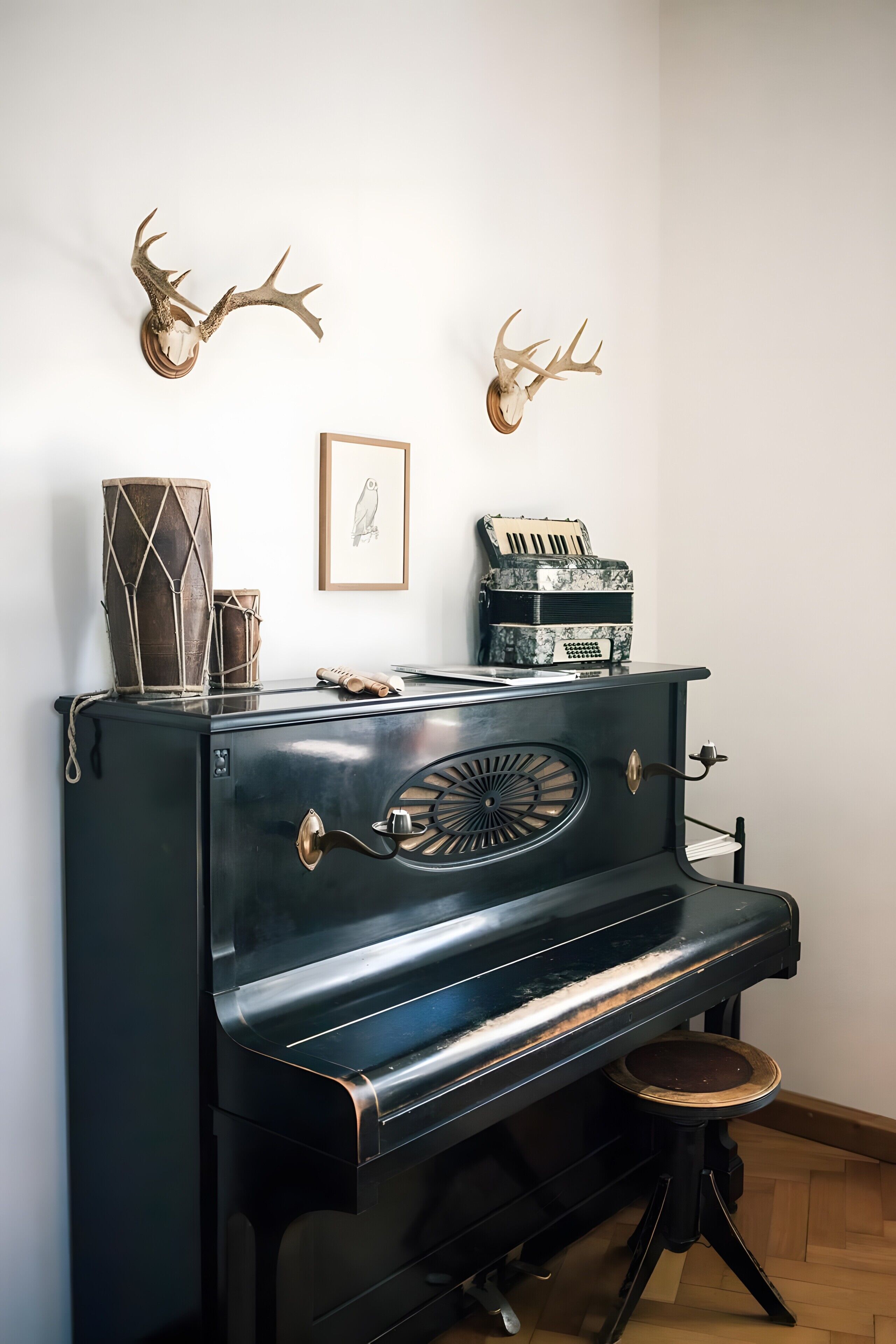A black piano with a stool, decorative antlers, a framed owl picture, and vintage radios on top.