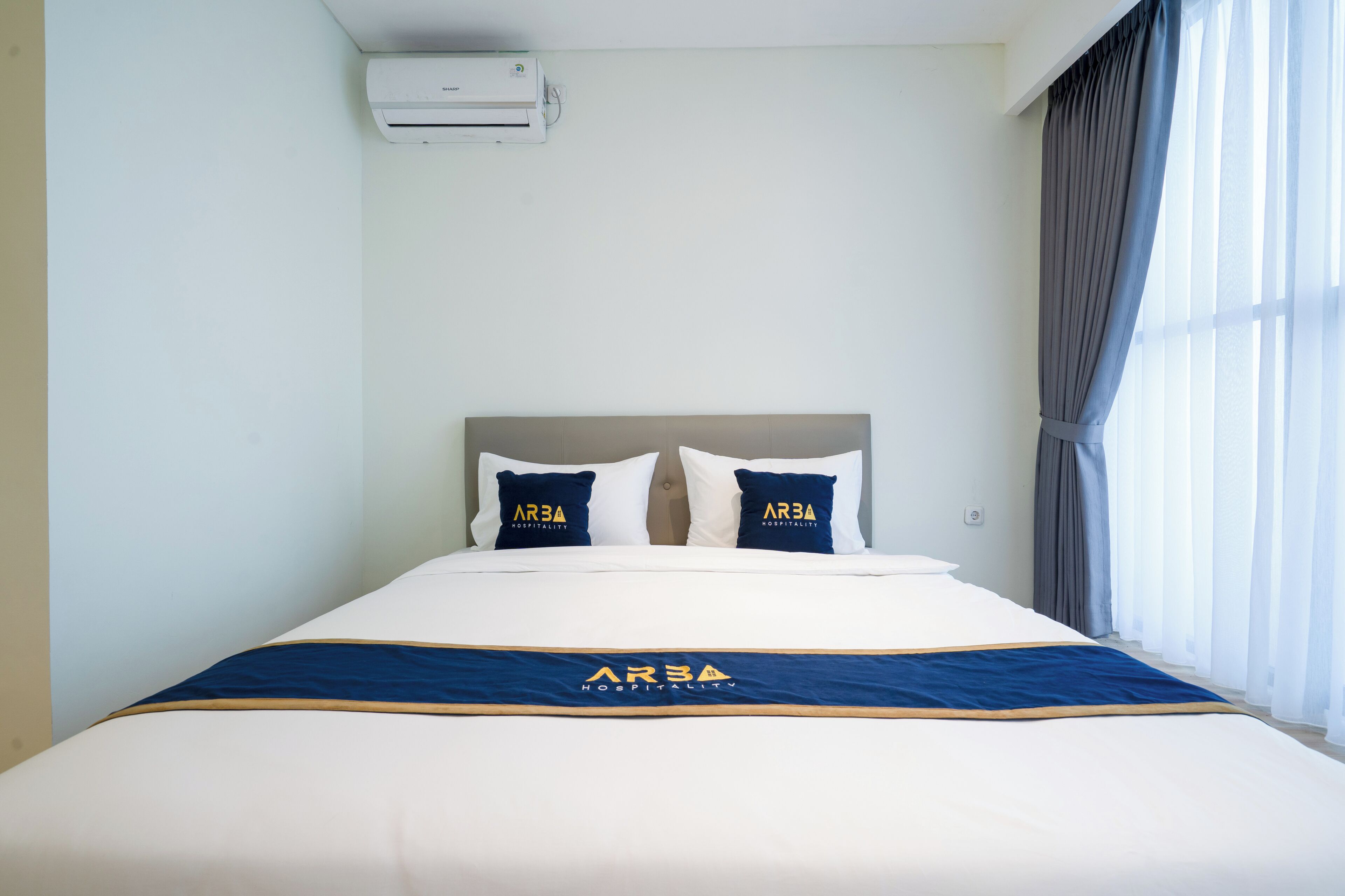 Een hotelkamer met een bed, twee kussens met 'ARBA'-branding, een blauw-gouden sprei en een raam met gordijnen.