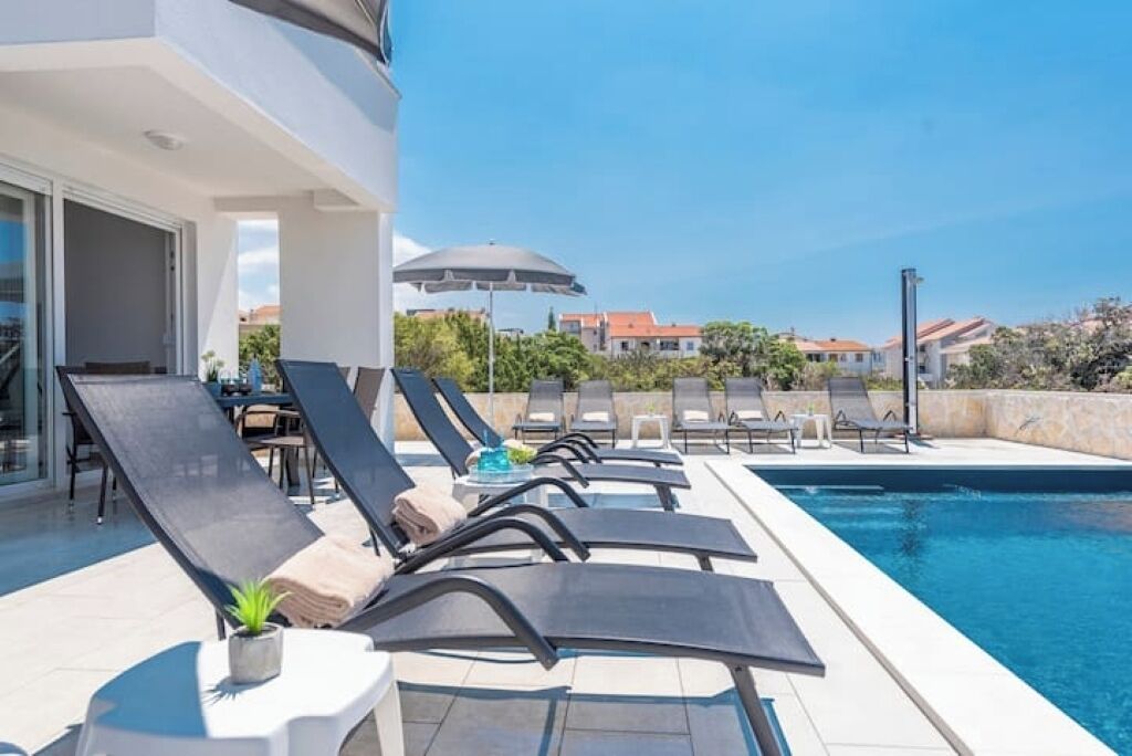 Ein Poolbereich mit Liegestühlen und Sonnenschirmen, ein klarer Himmel und Gebäude im Hintergrund.