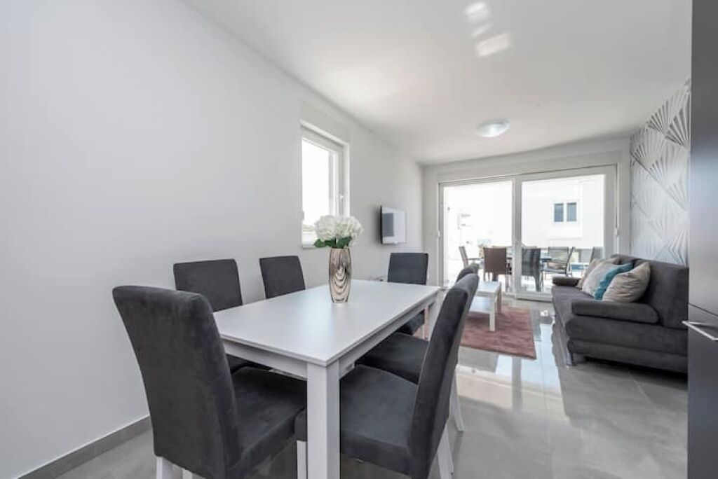 Een moderne woonkamer met een eethoek, met een witte tafel, grijze stoelen en een grijze bank.
