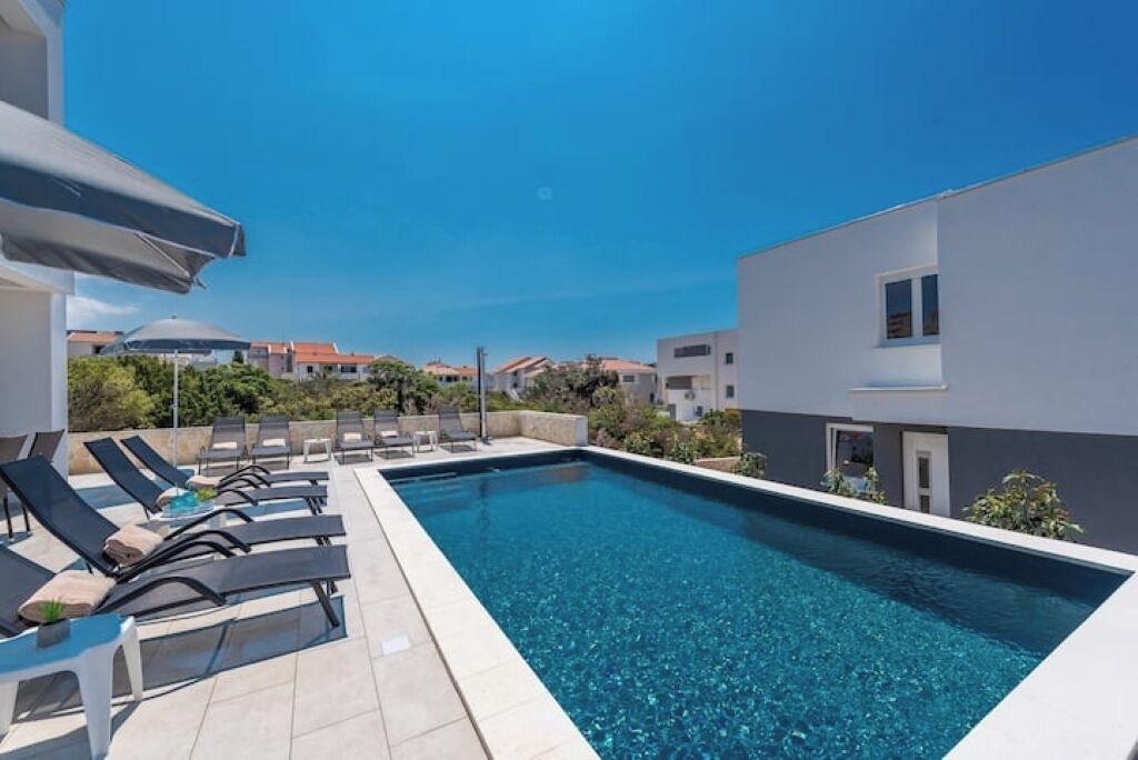 A rectangular swimming pool with clear blue water, surrounded by lounge chairs and a tiled deck.