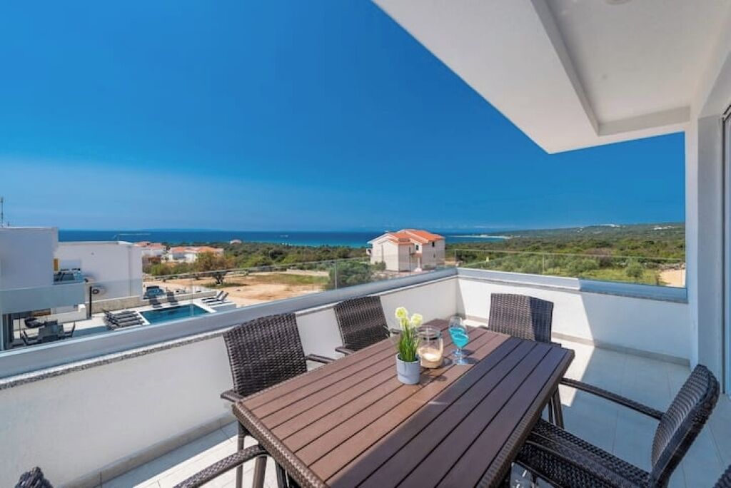 A balcony with a wooden table, wicker chairs, and a view of the sea and distant buildings.