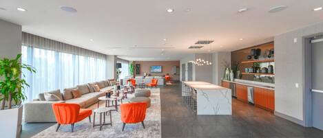 A spacious living area with a sofa, orange chairs, and a coffee table. There is a kitchen area with a bar and shelves with kitchenware.