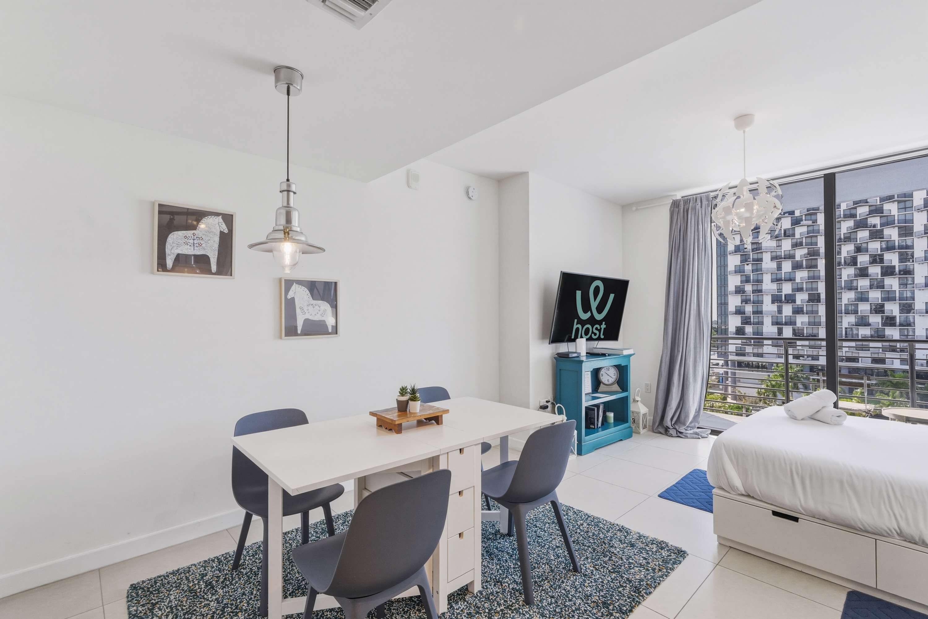 Quarto de hotel moderno com cama, área de refeições com mesa e cadeiras, televisão e uma grande janela com vista para a cidade.