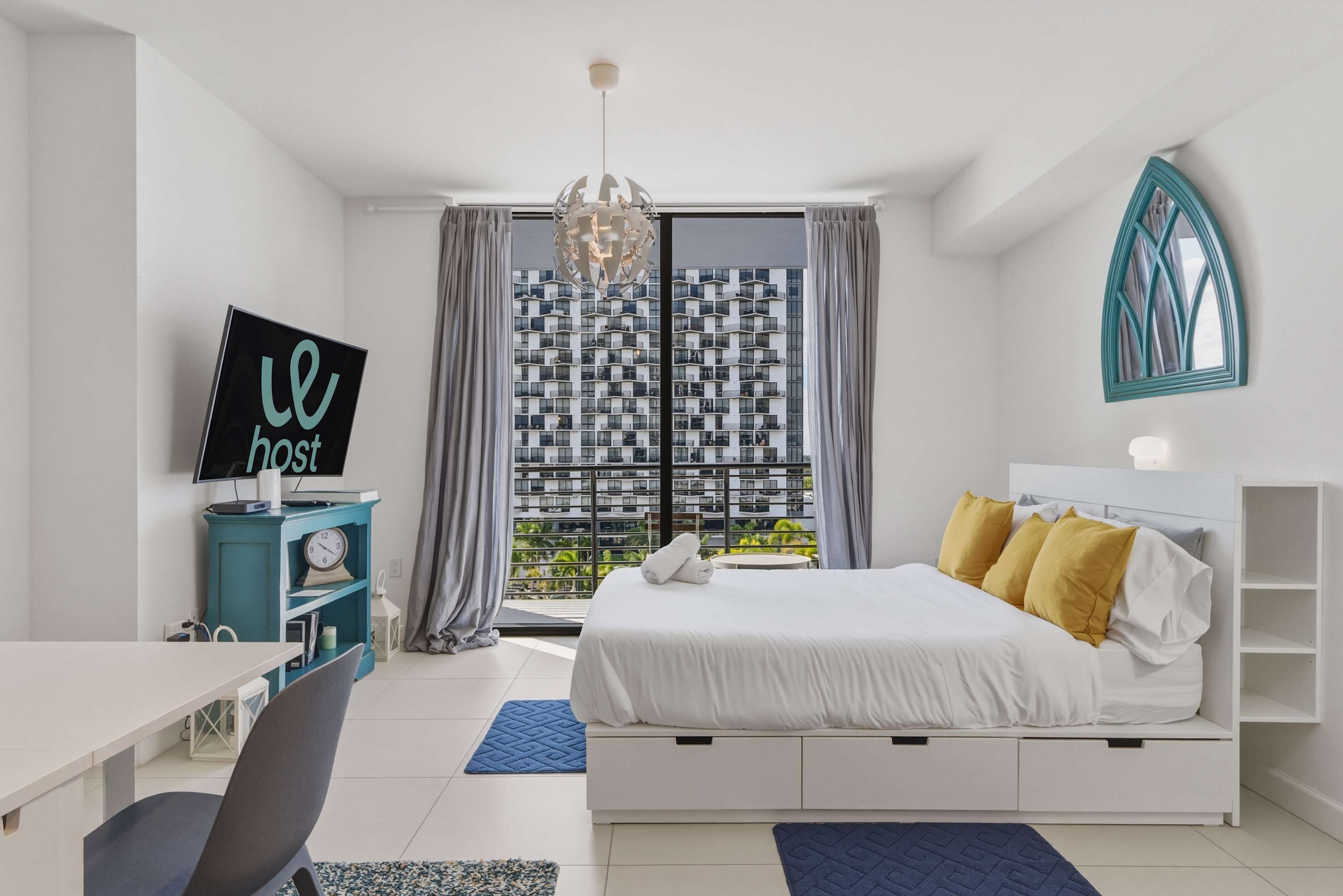 Een moderne hotelkamer met een bed, een bureau, een televisie en uitzicht op een stadsgezicht door grote ramen.