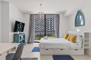 A modern hotel room with a bed, a desk, a TV, and a view of a cityscape through large windows.