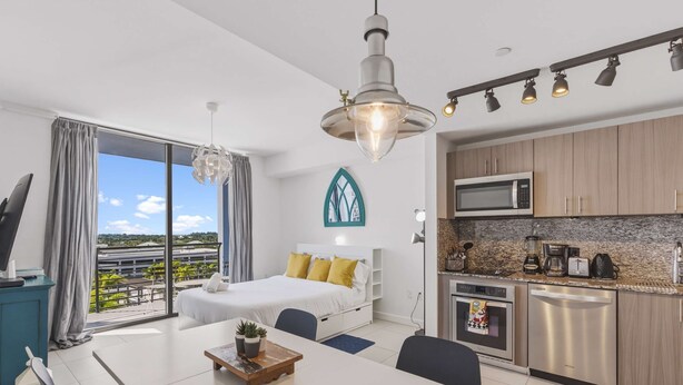 A modern hotel room with a balcony, a bed with yellow pillows, a dining area with a table and chairs, and a kitchenette with a microwave and toaster. - Studio in the Heart of Downtown Doral | Pool (Doral)