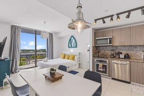 A modern hotel room with a balcony, a bed with yellow pillows, a dining area with a table and chairs, and a kitchenette with a microwave and toaster.