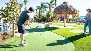 Uma pessoa jogando golfe num campo bem cuidado, com uma construção de colmo ao fundo.