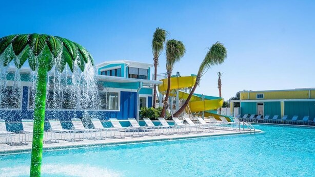 Uma piscina com elementos aquáticos, palmeiras e um edifício com cores azul e amarelo.