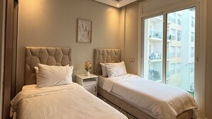 A modern hotel room with two beds, a small bedside table with a vase, and a large window offering a view of the city. - Appartement propre à Rabat – Location journalière (Rabat)