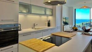 Een moderne keuken met een inbouwoven, een spoelbak en een groot eiland met een houten werkblad. Er zijn hangende lampen en uitzicht op de oceaan door grote ramen.