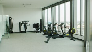 Une salle de sport équipée de divers appareils d’exercice, dotée de grandes fenêtres et offrant une vue sur la ville.