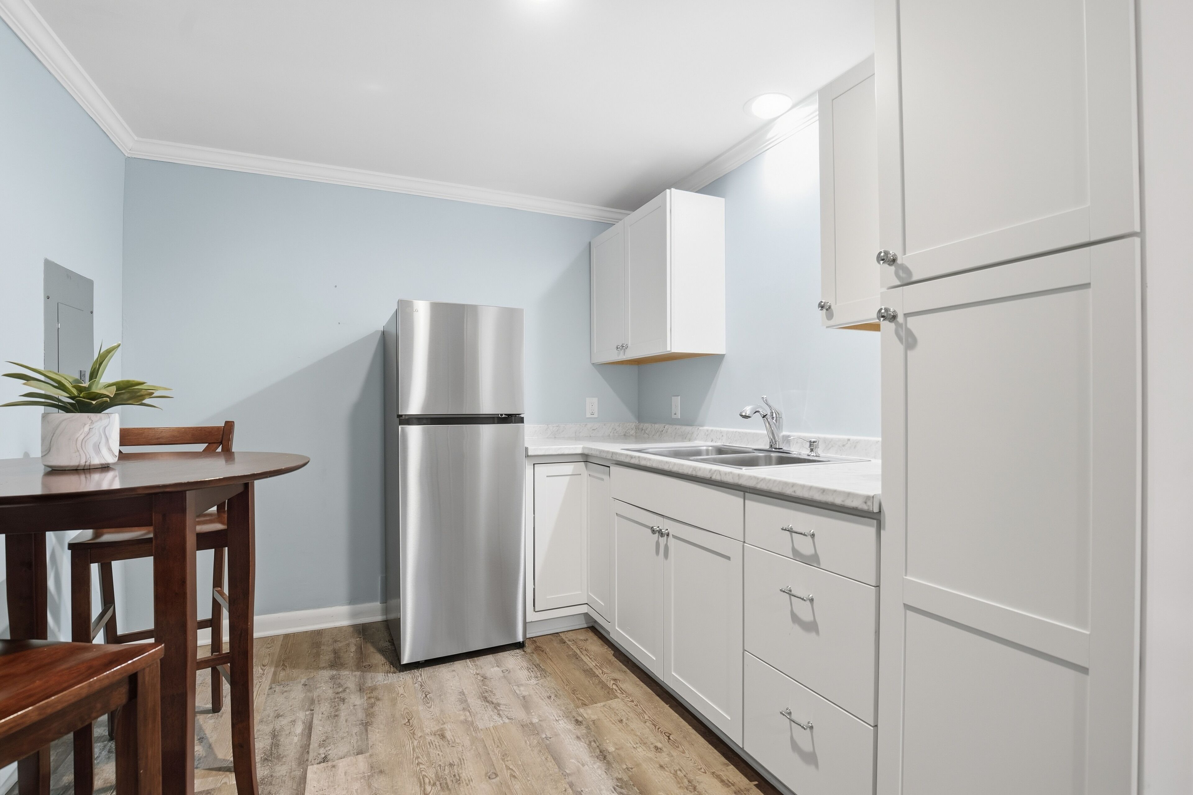 A kitchen with white cabinets, a stainless steel refrigerator, a wooden dining table, and a potted plant.